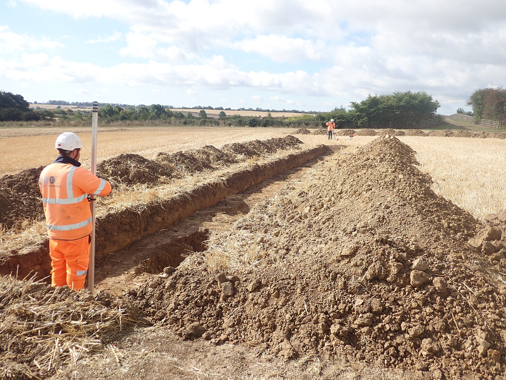 Two archaeologists surveying a long evaluation trench with a ranging rod and measuring equipment